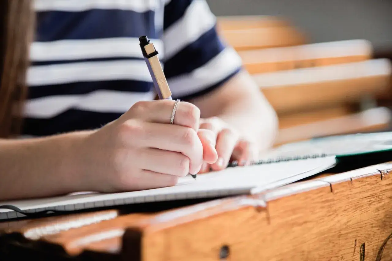 Dłoń w pierścionku pisze długopisem w zeszycie na drewnianej ławce.