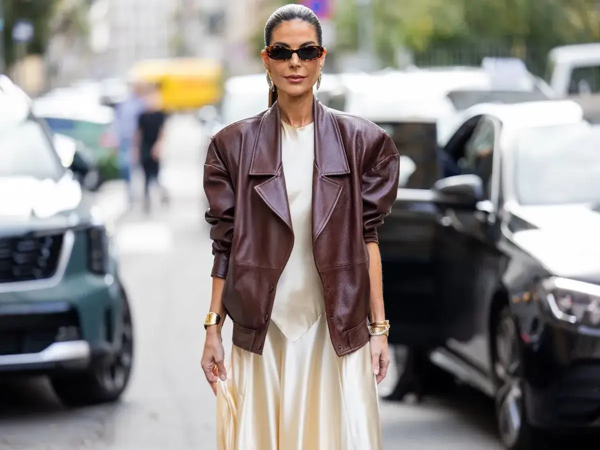 Kobieta w brązowej skórzanej kurtce i satynowej sukience stoi na ulicy.