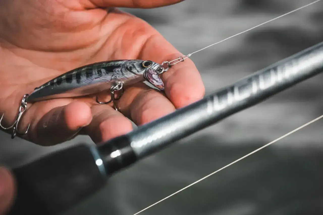 Dłoń trzymająca srebrnego woblera z haczykami i żyłką, obok wędki.