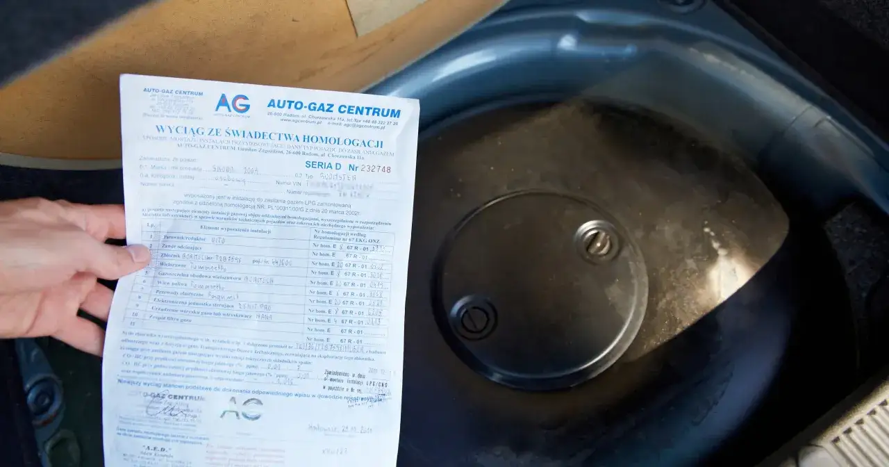 Fragment dokumentu z danymi homologacji butli LPG obok okrągłej pokrywy w bagażniku samochodu.