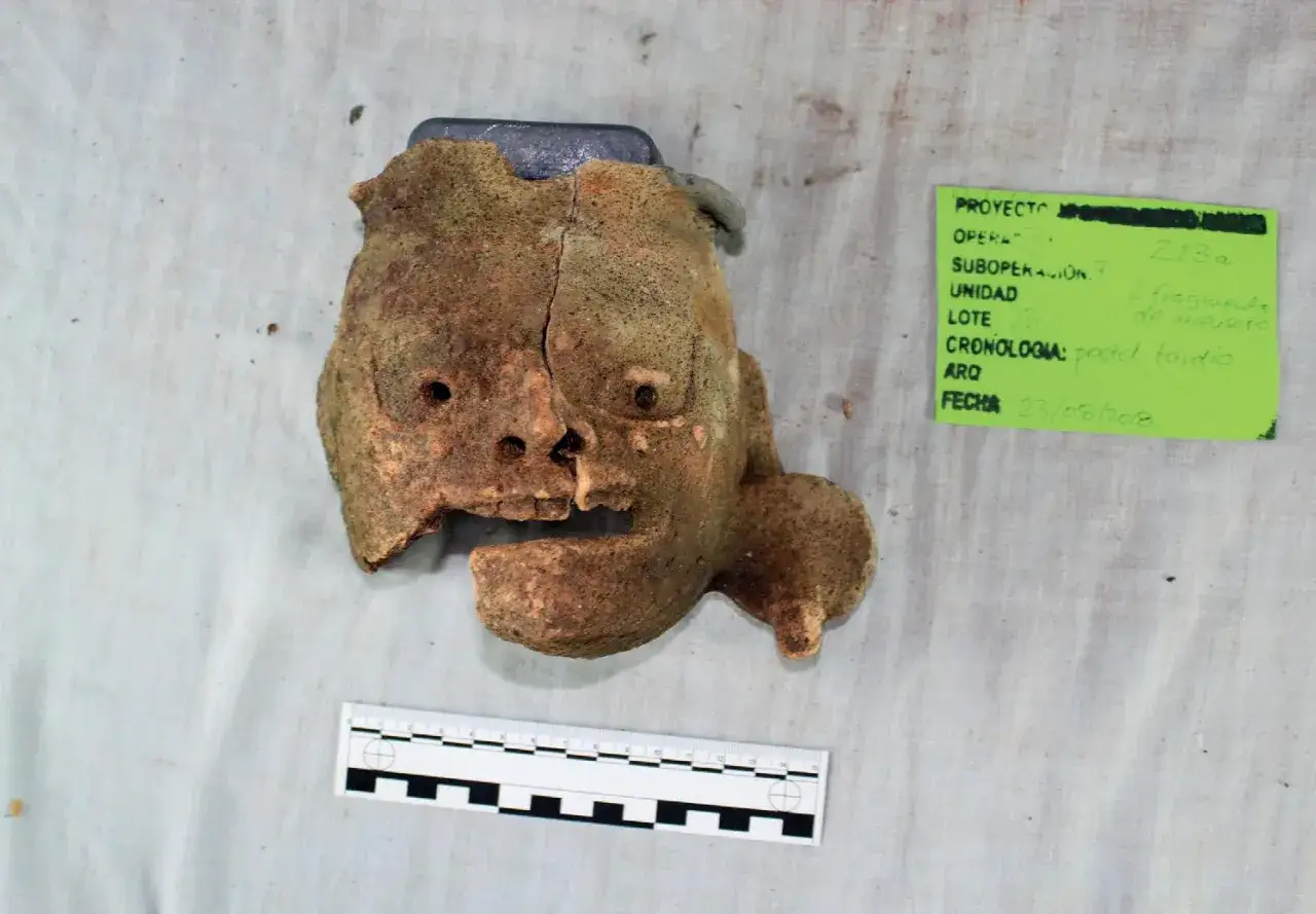 Fragment glinianej maski, jak z tekstu piosenki Kaen "Kim jestem?", z widocznymi otworami na oczy i usta, obok zielonej etykiety z datą 23/08/2018.