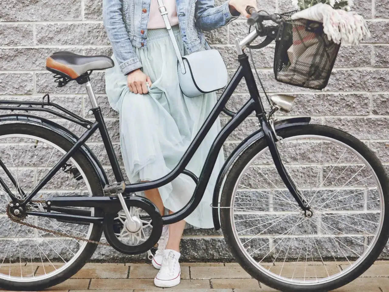 Elegancka kobieta w zwiewnej sukience i dżinsowej kurtce opiera się o rower. Widoczny rozmiar ramy roweru sugeruje, że jest to model miejski, idealny na przejażdżki po mieście.