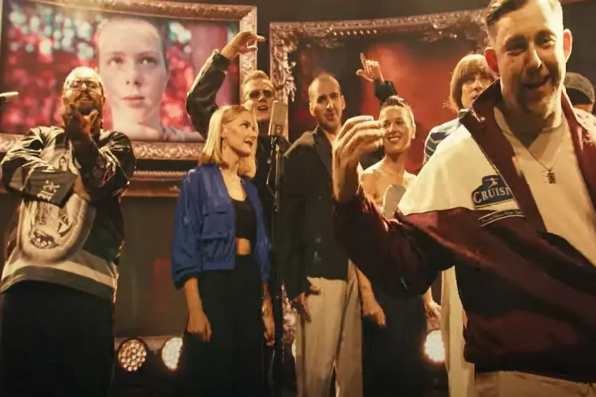 Grupa osób, w tym mężczyzna w bordowej kurtce, śpiewa "Witajcie w naszej bajce" na tle portretu.