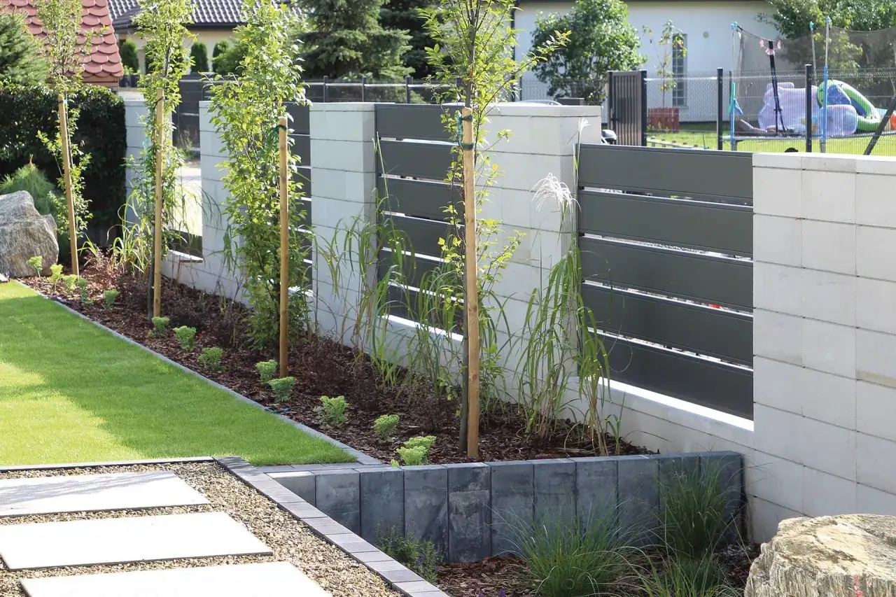 Nowoczesne ogrodzenie murowane pełne z betonowych bloczków i ciemnoszarych paneli, otaczające zadbany ogród z młodymi drzewkami.