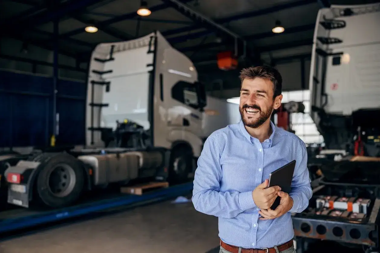 Uśmiechnięty mężczyzna z tabletem w ręku, w tle warsztat z ciężarówkami. Oferuje **usługę transportową ryczałt**.