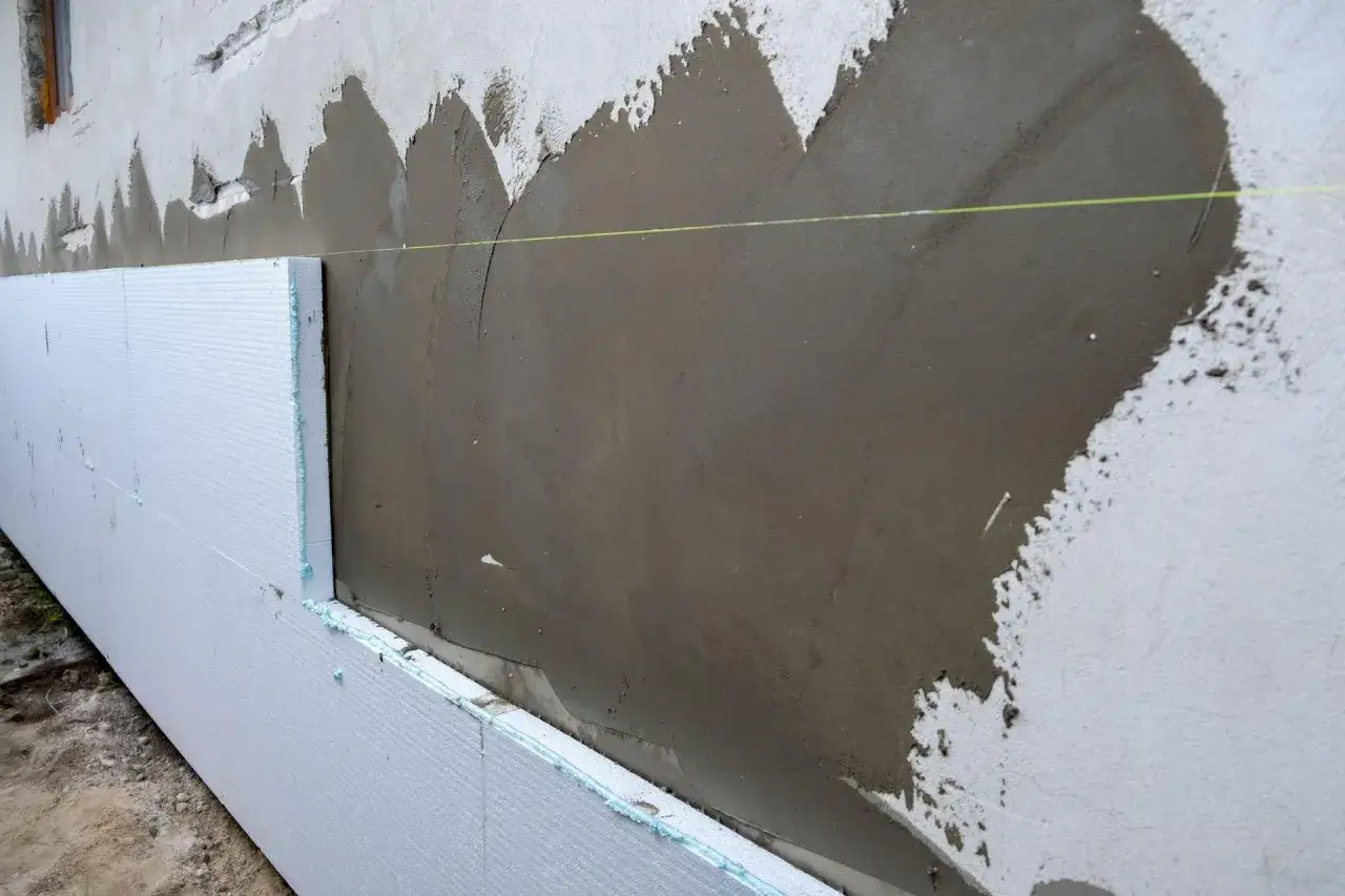 Ściana ze styropianu w trakcie ocieplania. Widać białe płyty styropianowe przyklejone do muru, na które nakładana jest szara zaprawa.