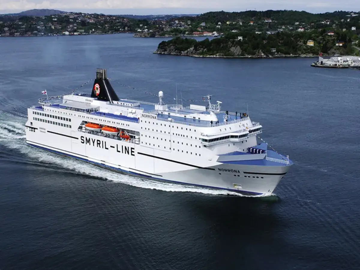 Duży biały prom "Smyril-Line Norrona" płynie po spokojnym morzu, w tle widać zielone wybrzeże.