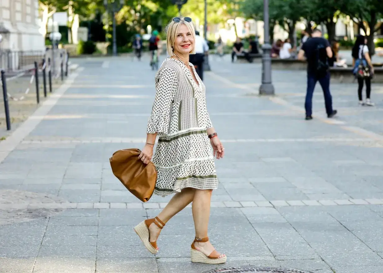 Kobieta w sukience w geometryczne wzory i espadrylach na koturnie idzie ulicą z brązową torebką.