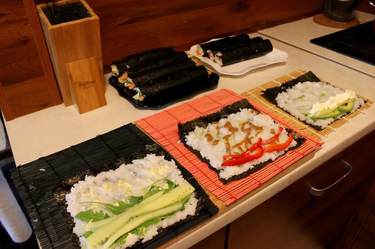 Przygotowanie ryżu do sushi. Na matach leżą arkusze nori z ryżem i dodatkami: ogórkiem, awokado, papryką i sosem.