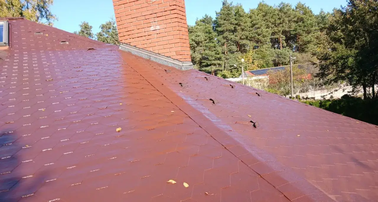 Czerwony dach pokryty gontem bitumicznym z widocznym kominem i drzewami w tle.