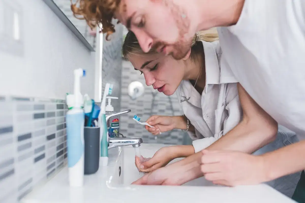 Para młoda nad umywalką. Ona płucze szczoteczkę, on pochyla się nad nią. Czy powinno się płukać zęby po umyciu?