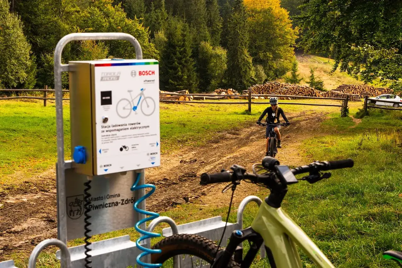 Stacje ładowania rowerów elektrycznych w Piwnicznej-Zdroju. W tle rowerzystka na szlaku.