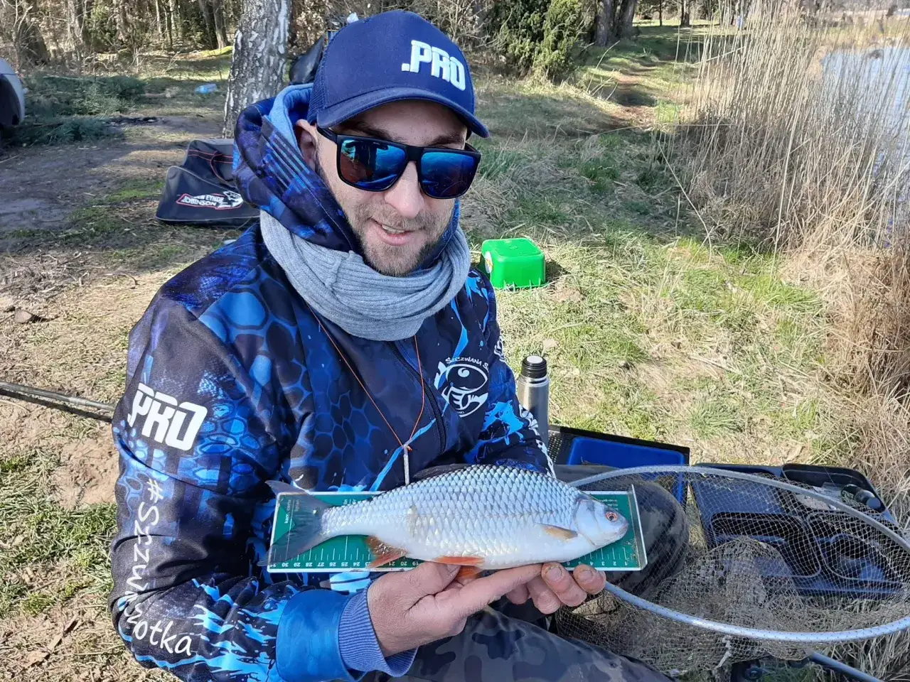 Wędkarz w niebieskiej czapce i bluzie z motywem PRO trzyma na miarce złowioną płoć.