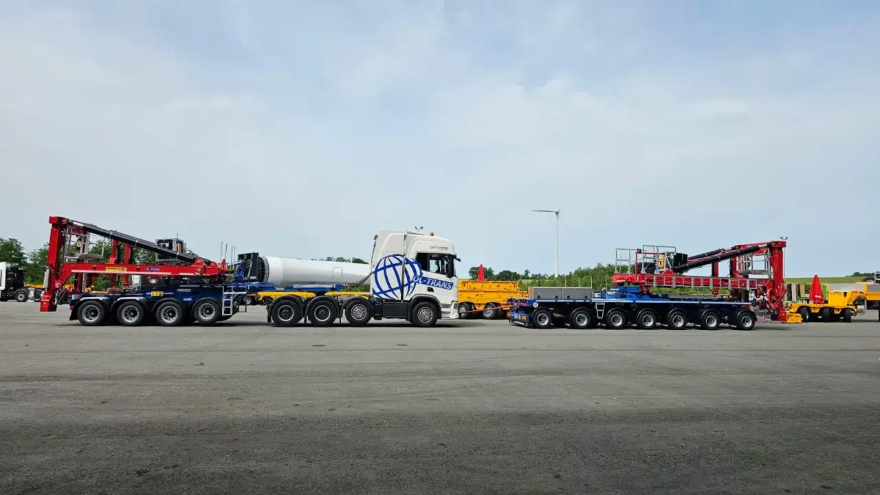 Ciężarówka z naczepą tandem przewozi element turbiny wiatrowej. Obok stoi inna naczepa z dźwigiem.