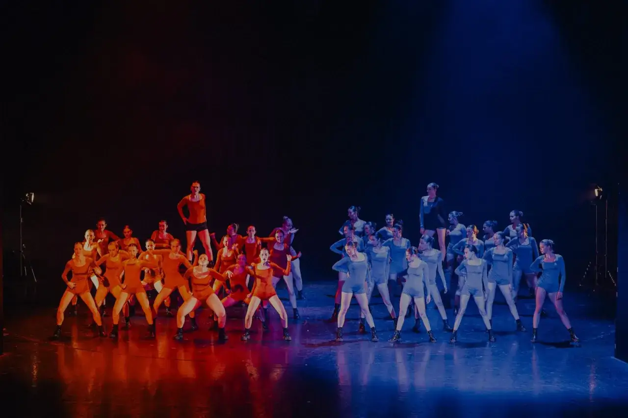 Grupa tancerzy na scenie, podzielona na dwie grupy: jedna w pomarańczowych strojach, druga w niebieskich. Nauka tańca nowoczesnego w pełnym blasku.