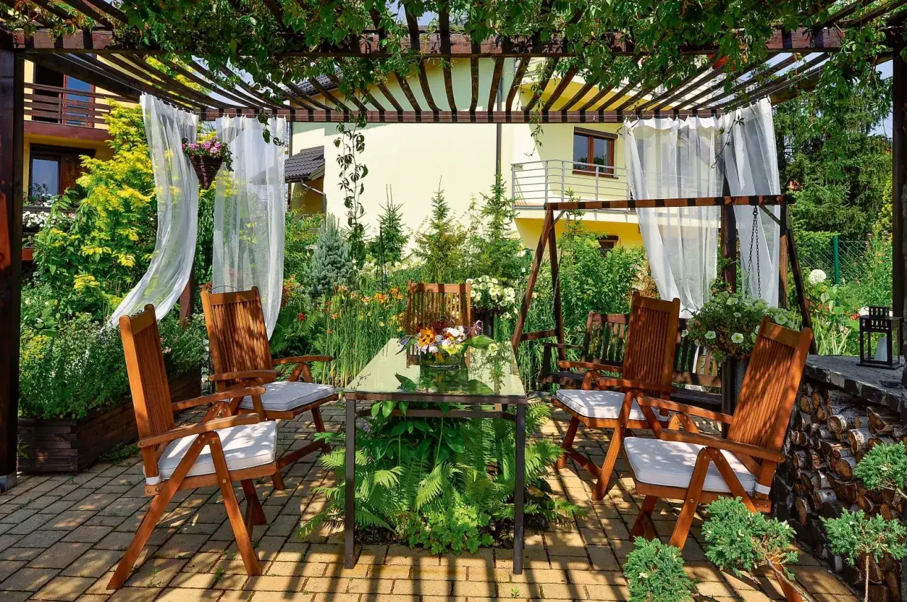 Urocze miejsce na relaks: drewniane meble, bujna zieleń i kwiaty pod pergolą. Idealne co na pergole, by cieszyć się latem.