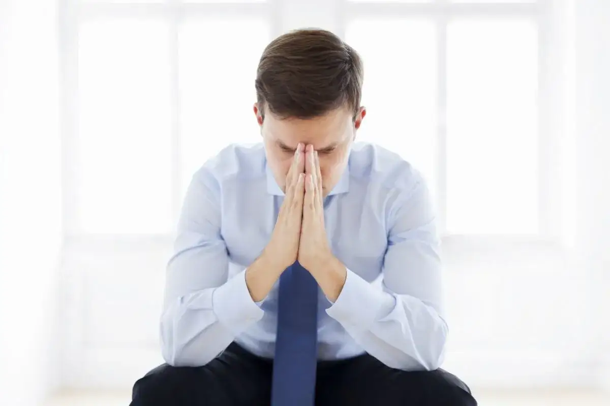 Mężczyzna w koszuli i krawacie z rękami złożonymi na twarzy, w zamyśleniu.