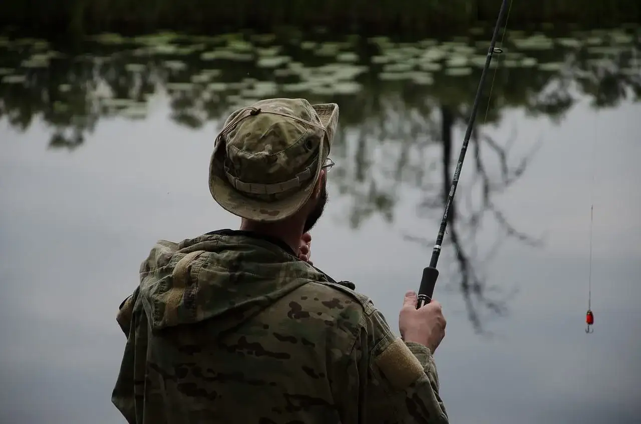 Mężczyzna w kamuflażu przygotowuje się do łowienia karpia. Wędka z haczykiem i przynętą w wodzie.