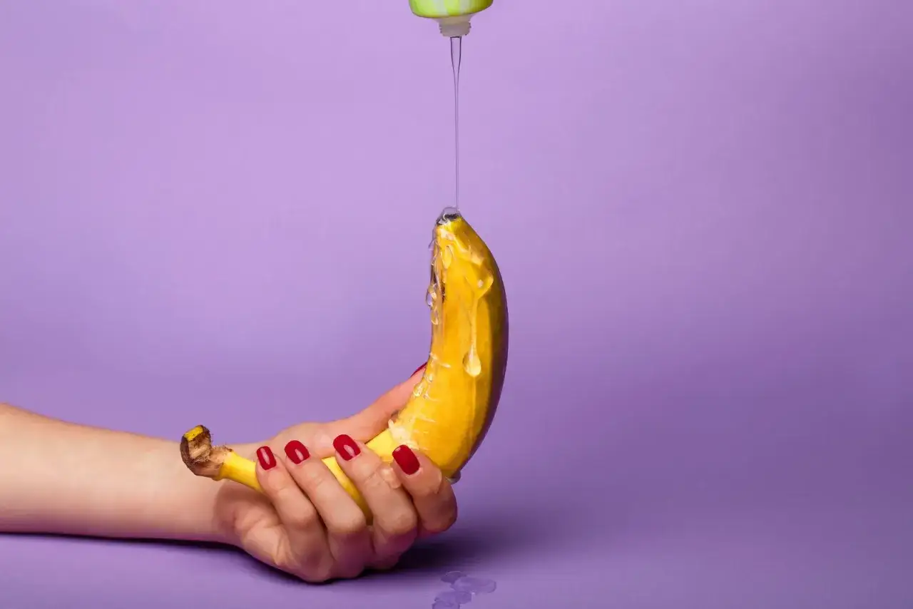 Dłoń z czerwonymi paznokciami trzyma banana, na który kapie przezroczysty lubrykant. Jaki lubrykant do prezerwatywy?