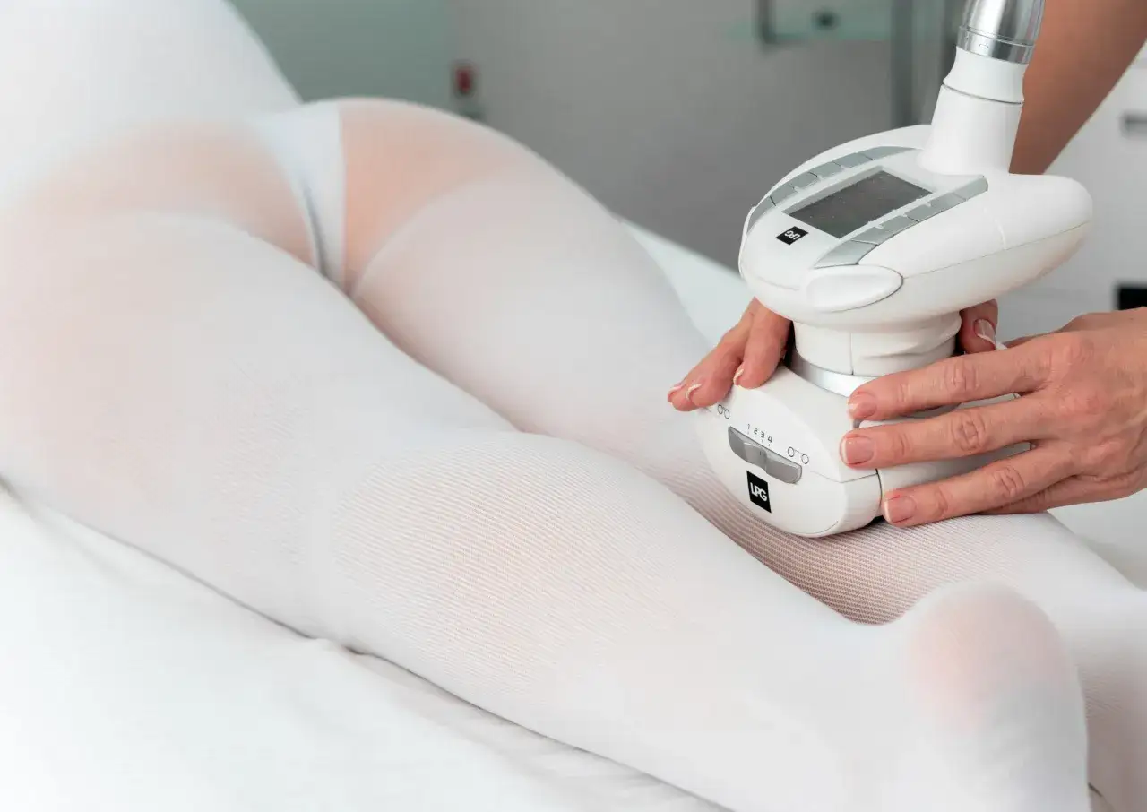 Zabieg endermologii w domu. Specjalistka wykonuje masaż specjalistyczną głowicą na nogach pacjentki w białej bieliźnie.