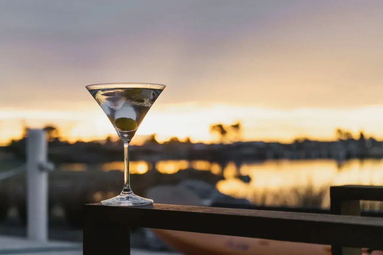 Idealny martini wstrząśnięte nie mieszane, z oliwką, na tle zachodzącego słońca nad wodą.