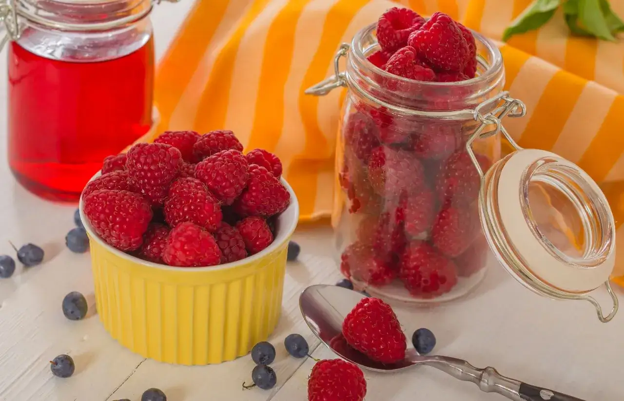 Sok z malin przepis babci: świeże maliny w żółtej miseczce i słoiku, obok czerwony napój i borówki.
