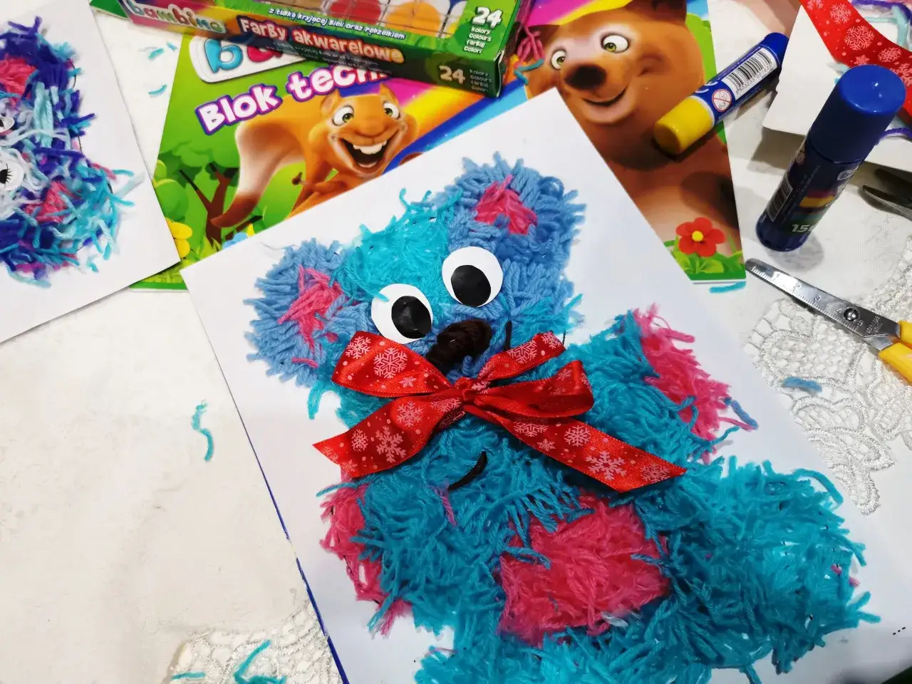 Pluszowy miś z włóczki w kolorach niebieskim i różowym, z czerwoną wstążką.