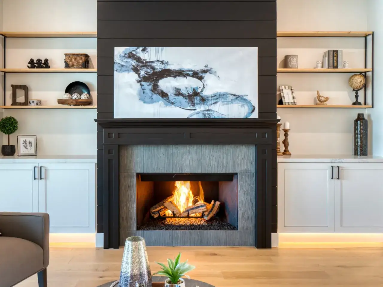 Nowoczesny kominek z palącym się ogniem, obrazem abstrakcyjnym i półkami z dekoracjami.