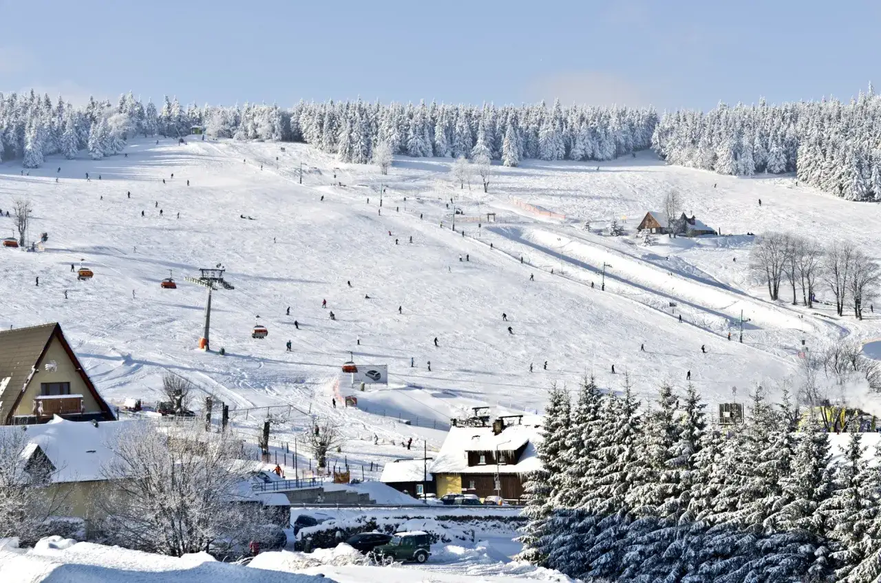 Ośnieżony stok narciarski z narciarzami, wyciągami i budynkami w tle.
