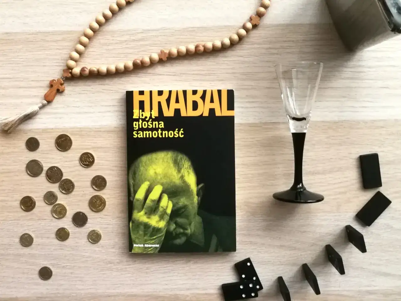 Książka Bohumila Hrabala "Zbyt głośna samotność", paciorki różańca, monety i kostki do gry na drewnianym blacie.