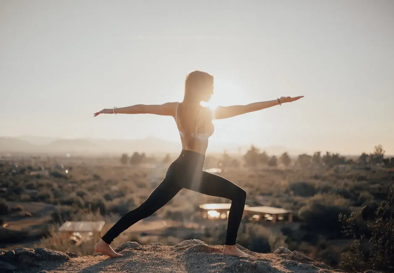 Kobieta w pozie wojownika na skale, szukająca spokoju. Słońce zachodzi, tworząc ciepłą poświatę.