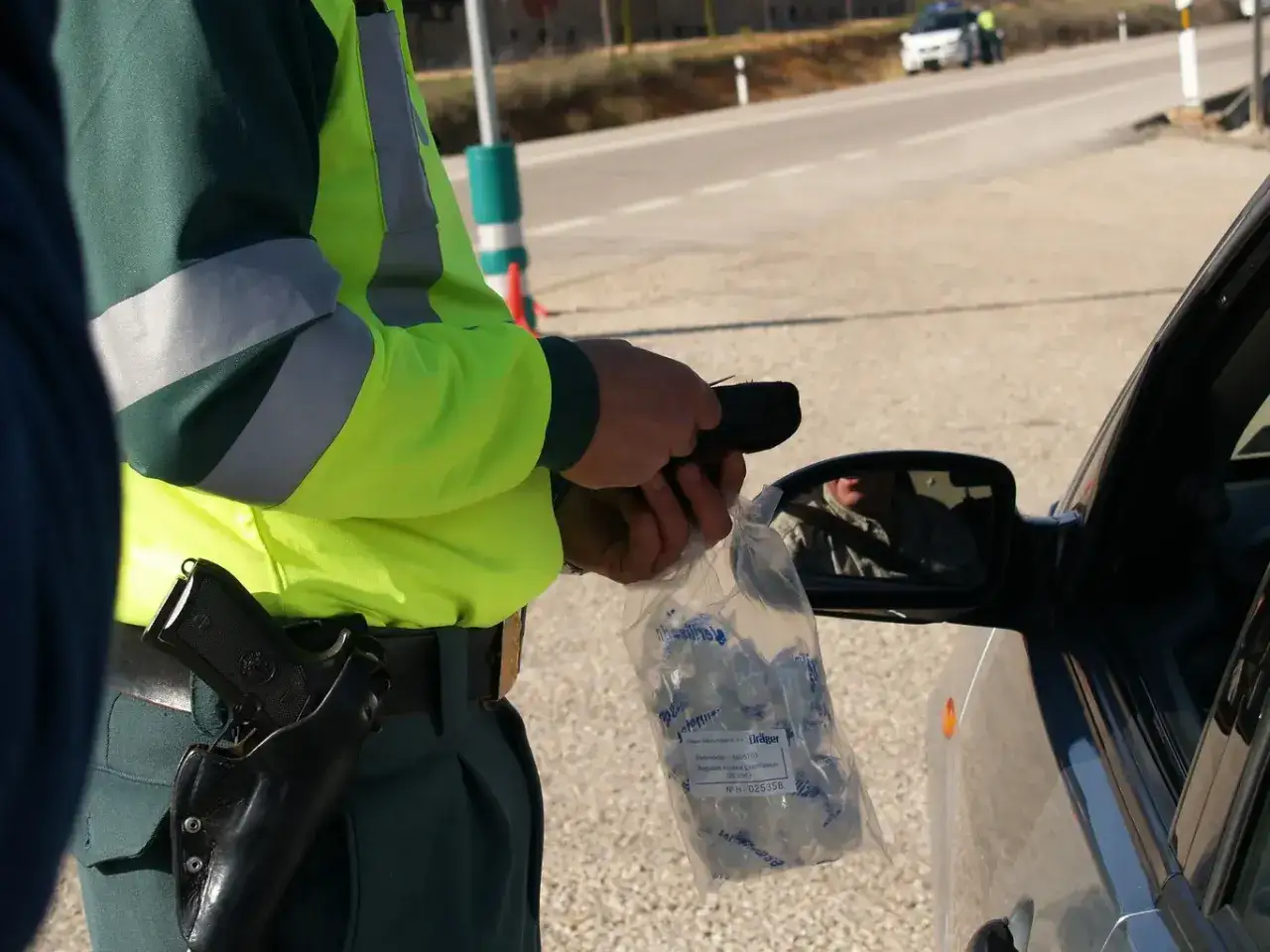 Policjant w kamizelce odblaskowej trzyma alkomat i worek z ustnikami przy samochodzie.