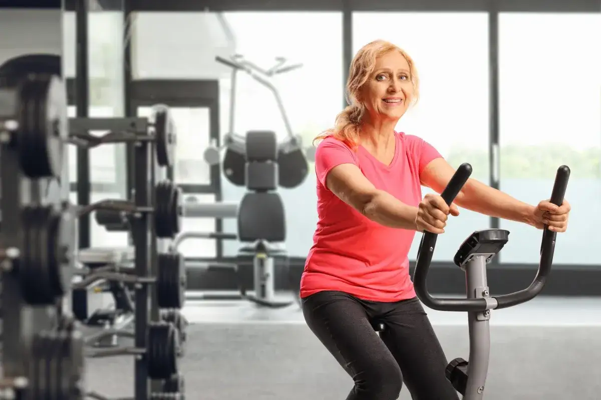 Uśmiechnięta starsza kobieta ćwiczy na rowerze stacjonarnym. Dobry puls podczas jazdy na rowerze stacjonarnym to klucz do efektywnego treningu.