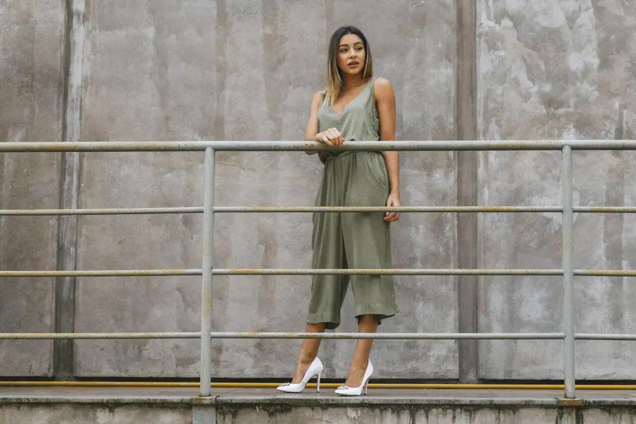 Kobieta w zielonym kombinezonie i białych szpilkach stoi przy metalowej balustradzie na tle betonowej ściany.