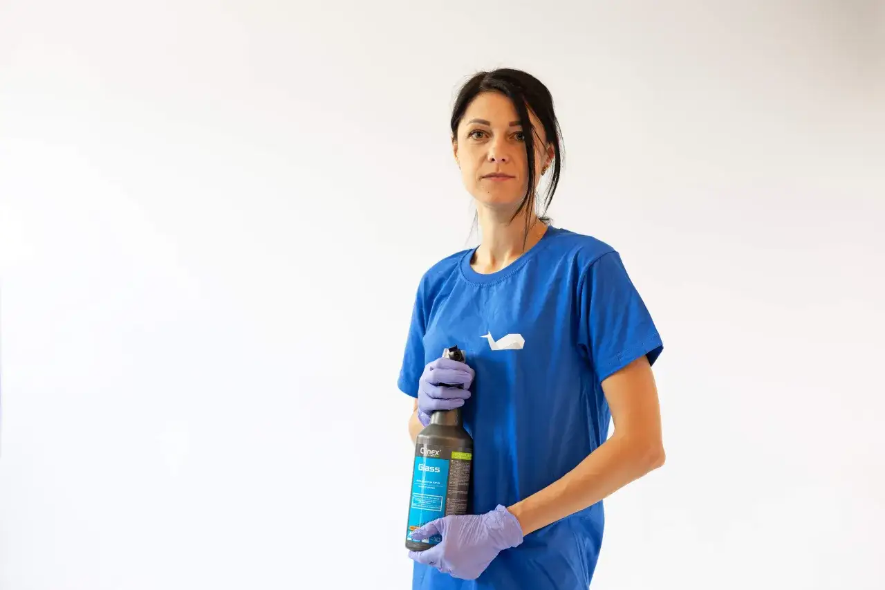 Kobieta w niebieskiej koszulce i fioletowych rękawiczkach trzyma butelkę płynu do mycia szyb.