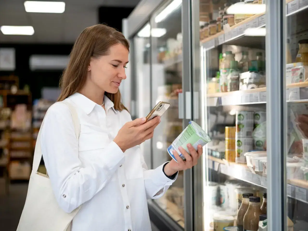 Kobieta w białej koszuli trzyma jogurt i telefon, analizując produkt w sklepie.
