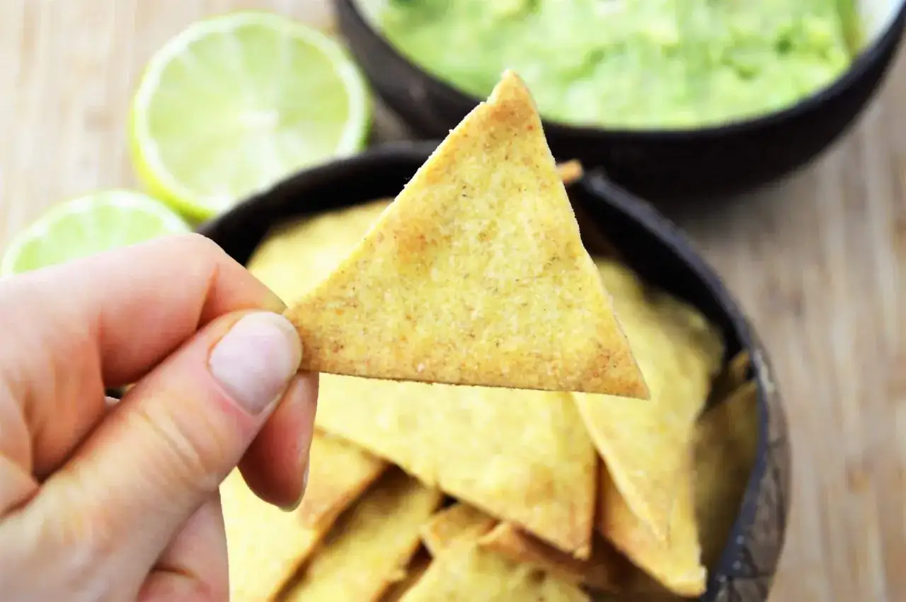 Dłoń trzyma nachos nad miską z guacamole, obok połówki limonki.