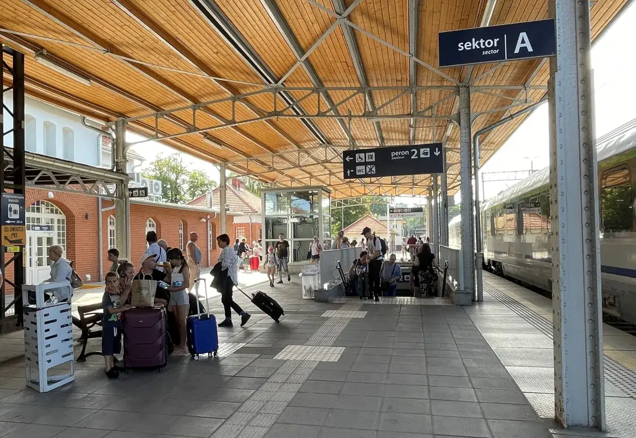 Ludzie z bagażami czekają na peronie. Znak "sektor A" i "peron 2" sugerują, że to stacja kolejowa, gdzie można dowiedzieć się, jak dojechać do Kołobrzegu pociągiem.