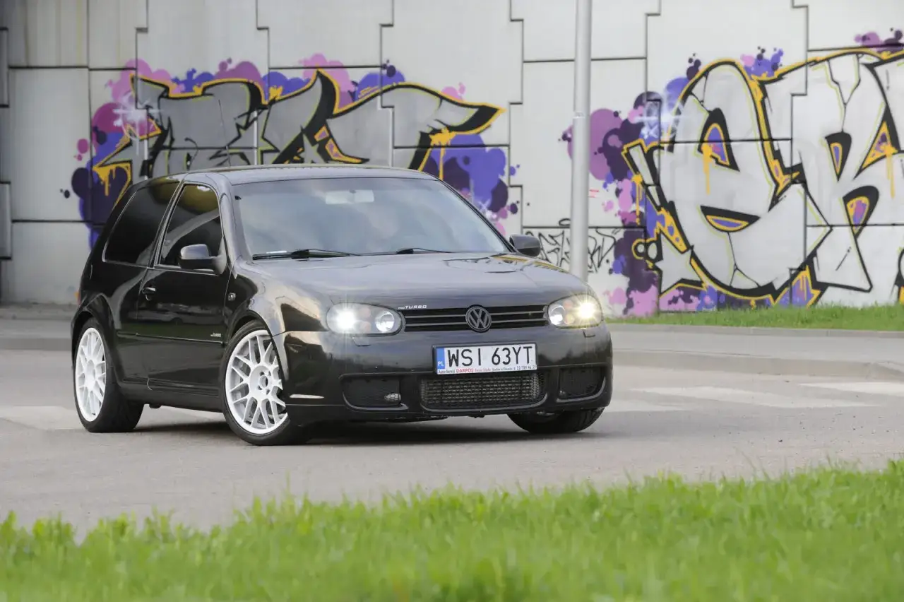 Czarny Golf 4 z tuningowym body kitem i białymi felgami, zaparkowany na tle kolorowego graffiti.