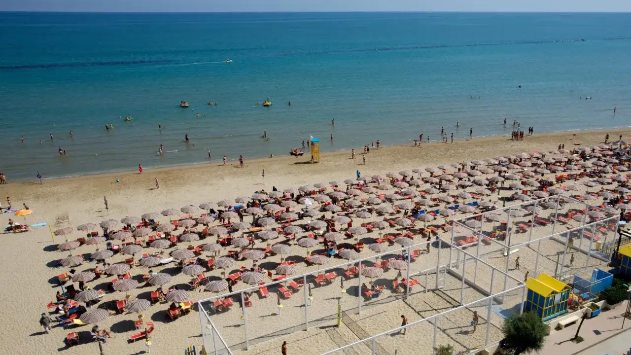 A crowded beach with rows of umbrellas and sunbathers, and people swimming in the blue sea.