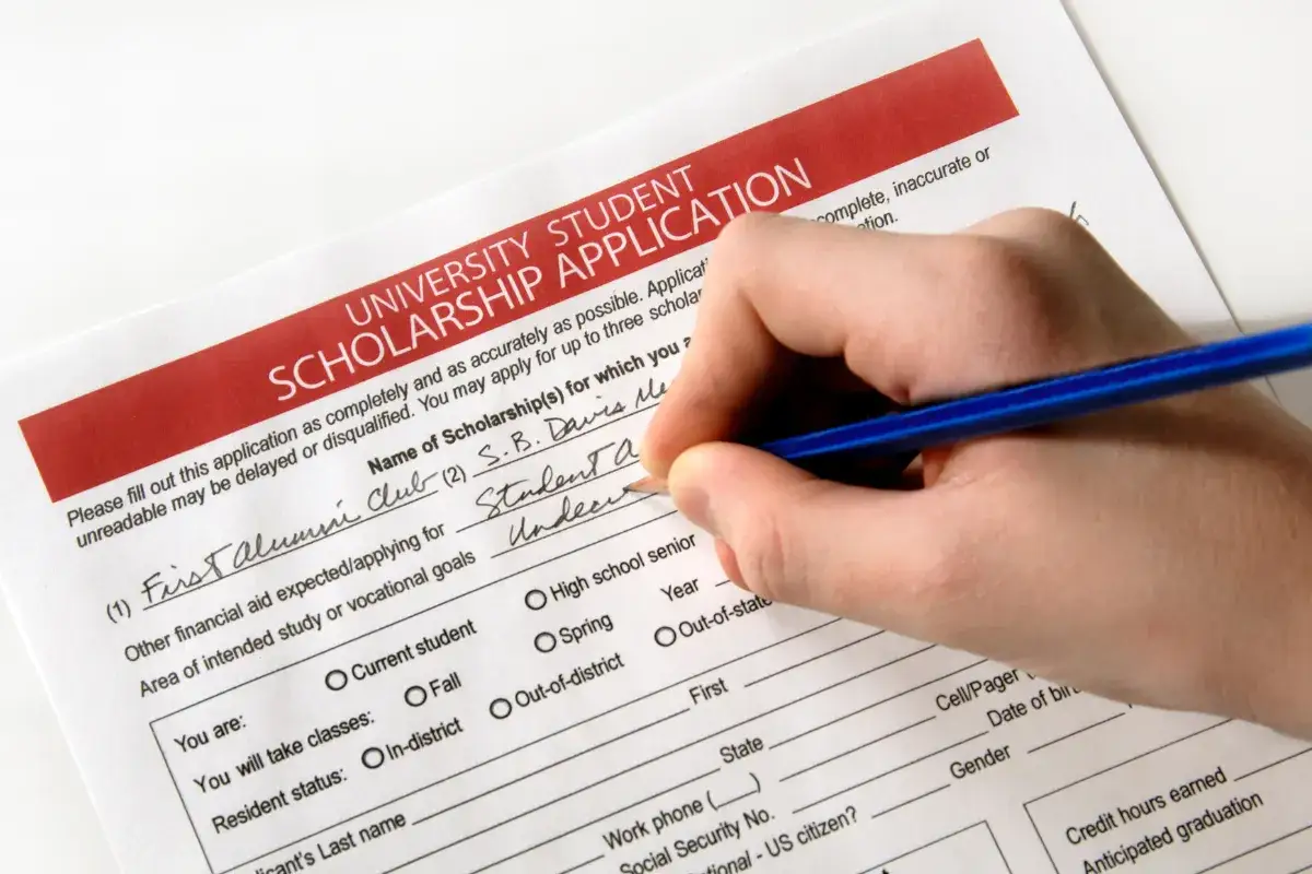 Dłoń z niebieskim ołówkiem wypełnia wniosek o stypendium uniwersyteckie.