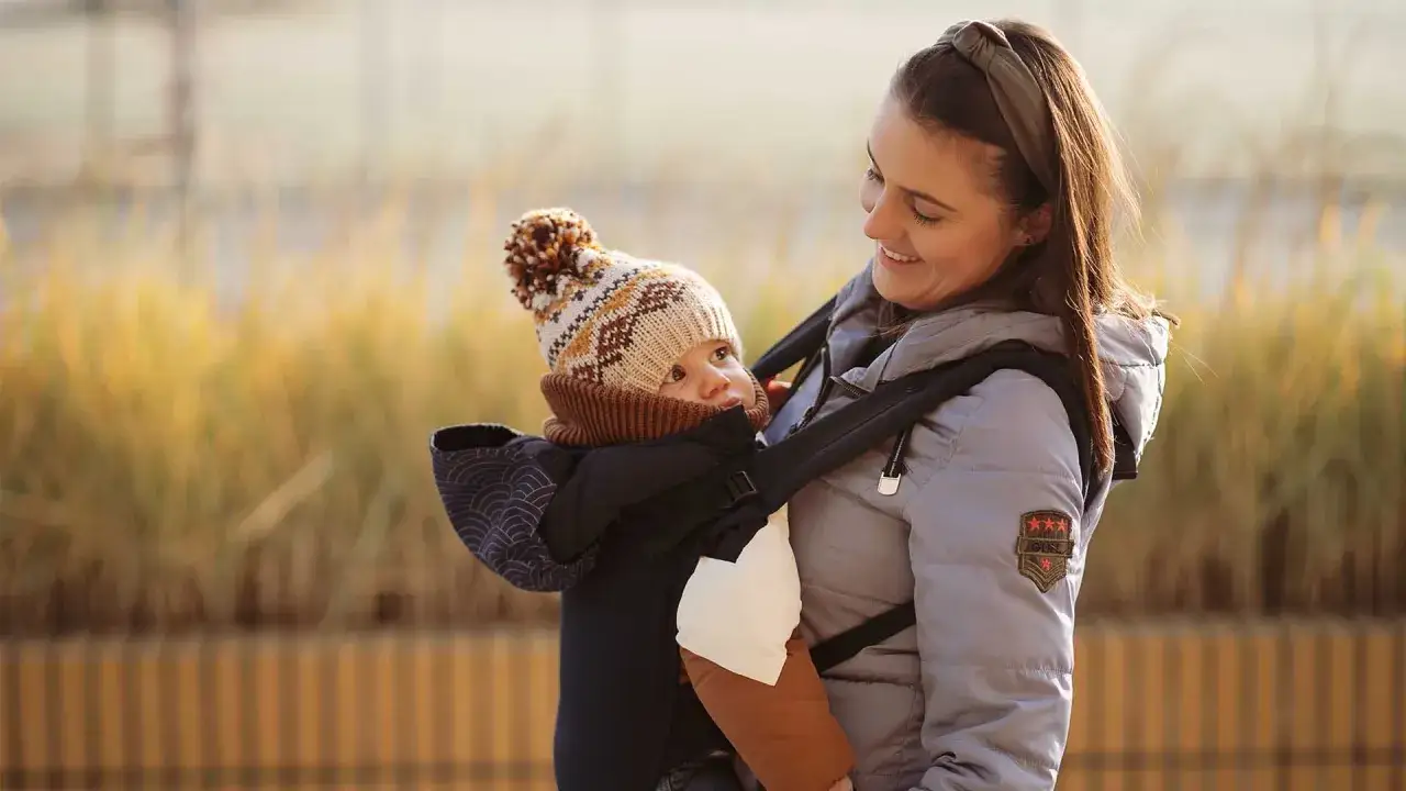 Fizjoterapeuta poleca nosidełko dla niemowlaka, które zapewnia prawidłową pozycję bioder i kręgosłupa. Uśmiechnięta mama z dzieckiem w nosidełku.