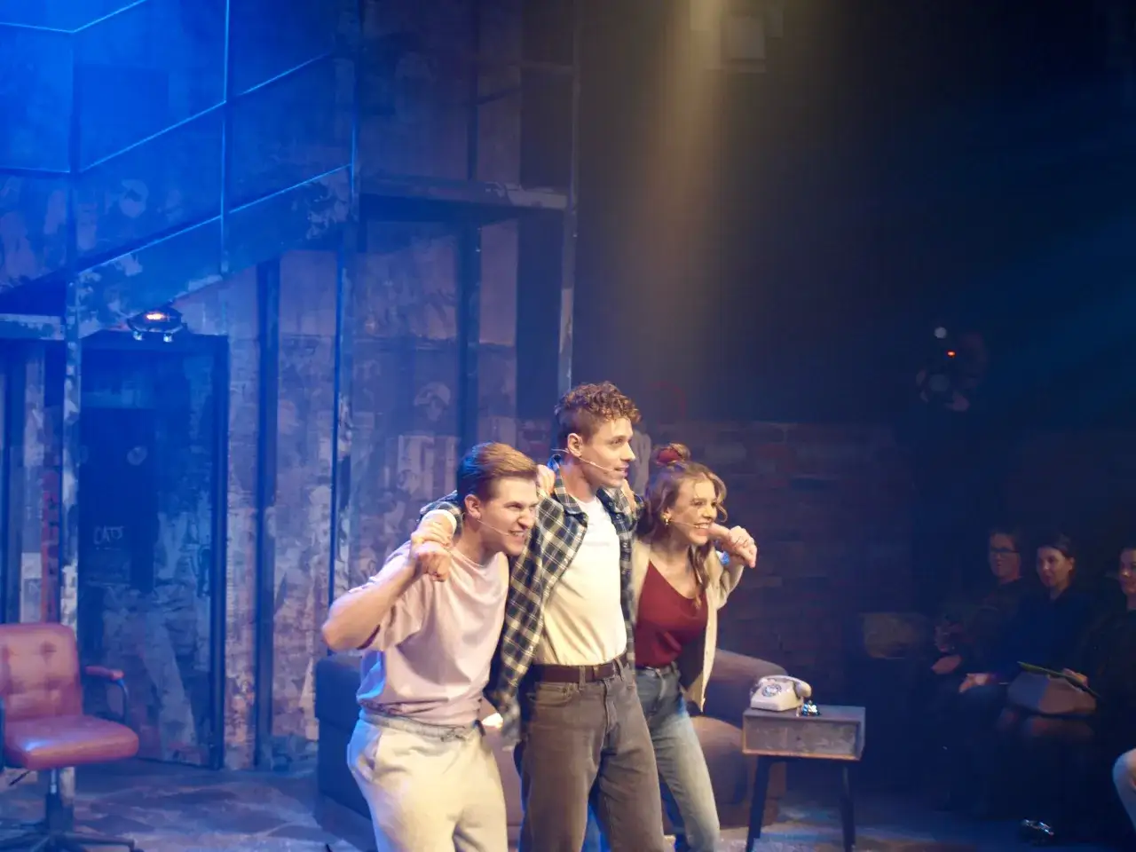 Trzech młodych ludzi na scenie, w tle ceglana ściana i niebieskie światła. Może to scena z musicalu "Tik Tik Bum" w Rzymie?