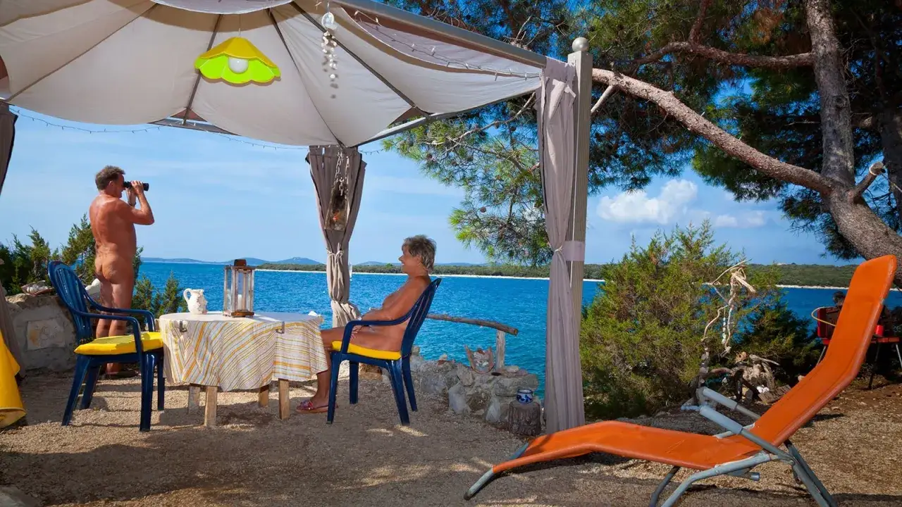 Para na plaży, relaksująca się w namiocie. Mężczyzna obserwuje morze przez lornetkę. To idealne miejsce na naturystyczny camping.