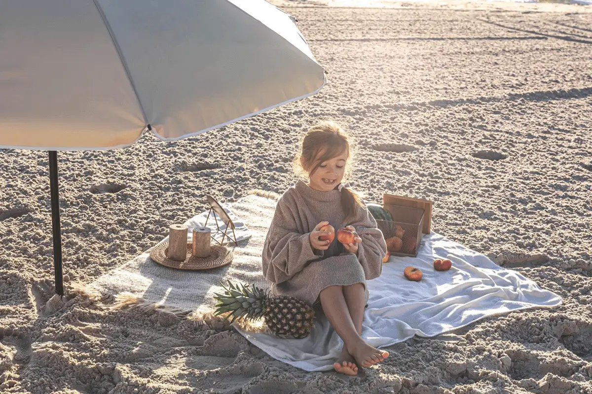 Dziewczynka na plaży pod parasolem, jedząca owoce. Idealny zestaw na wakacje nad morze z dzieckiem: koc, owoce, ananas.