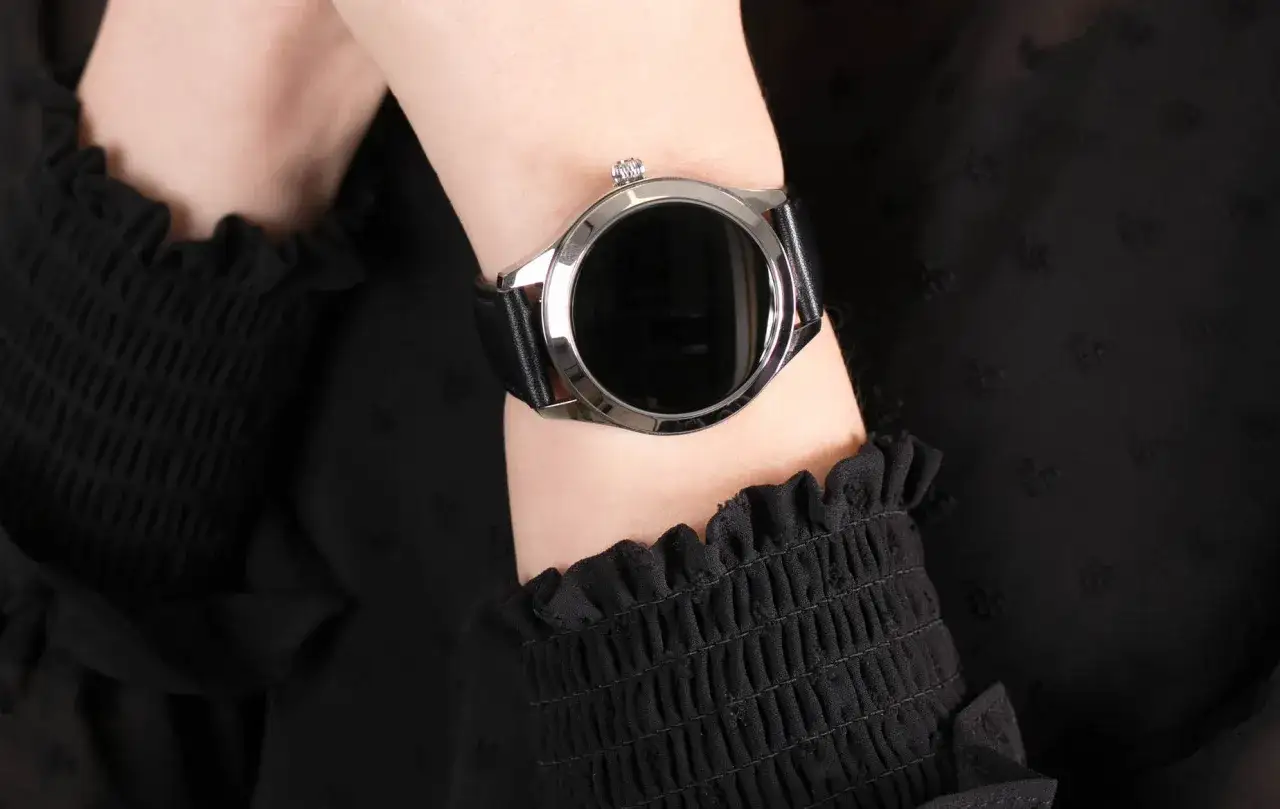 Damski smartwatch z czarnym paskiem na nadgarstku, na tle czarnej bluzki.