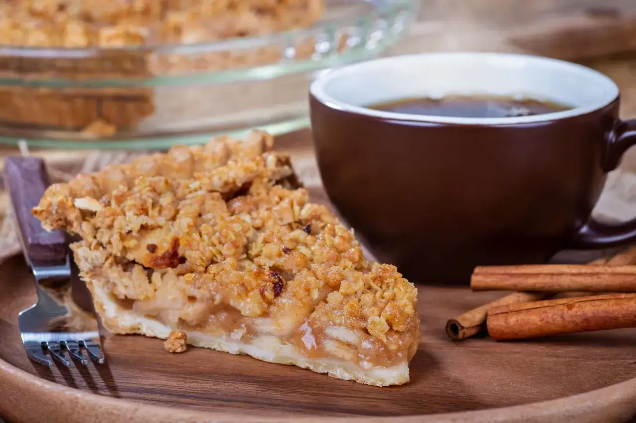 Kawałek jabłecznika z kruszonką, filiżanka kawy i laski cynamonu na drewnianej desce.