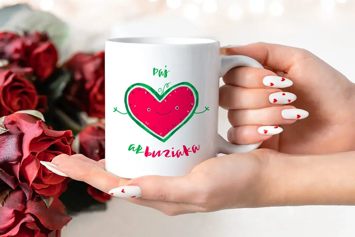 Biały kubek z zabawnym sercem-arbuzem i napisem "Daj buziaka", trzymany w dłoniach z paznokciami w serduszka, na tle czerwonych róż.