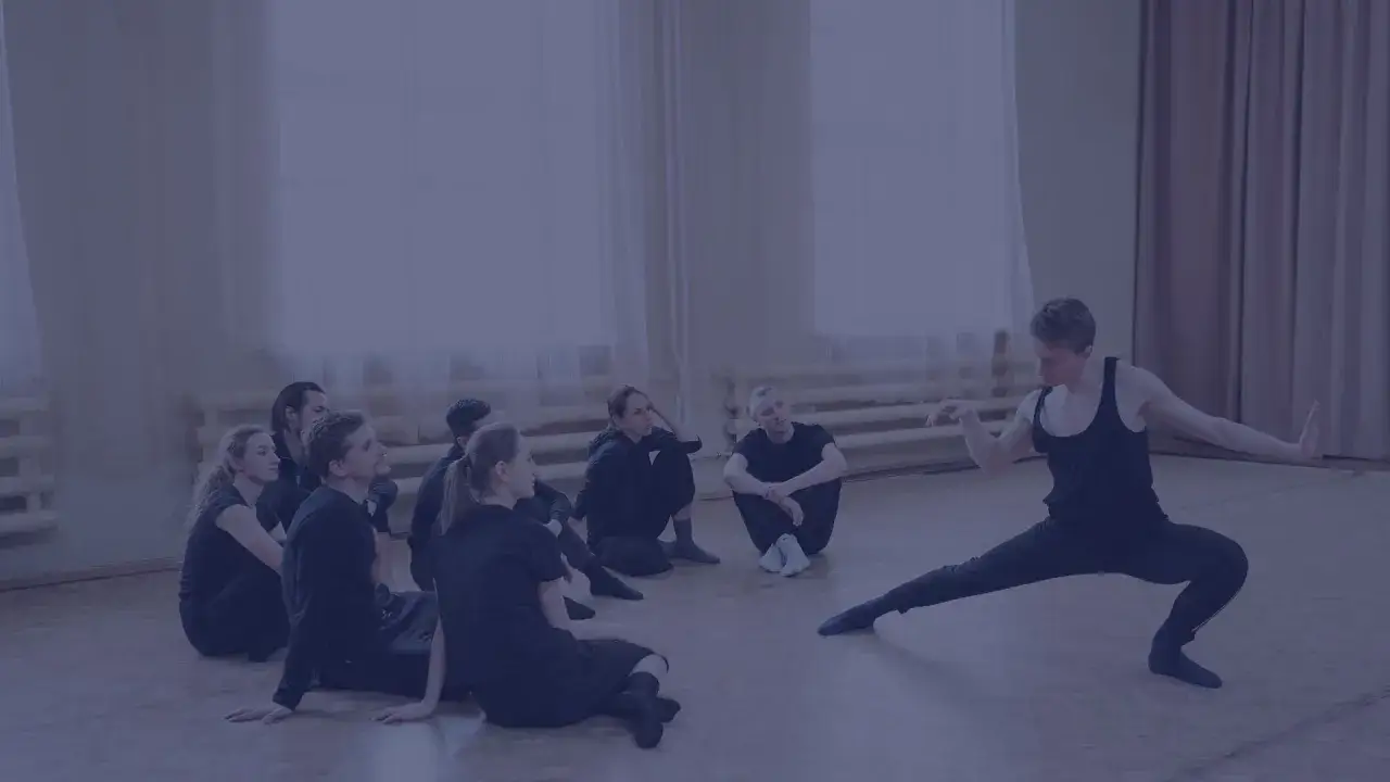 Choreograf demonstruje ruchy grupie tancerzy siedzących na podłodze.