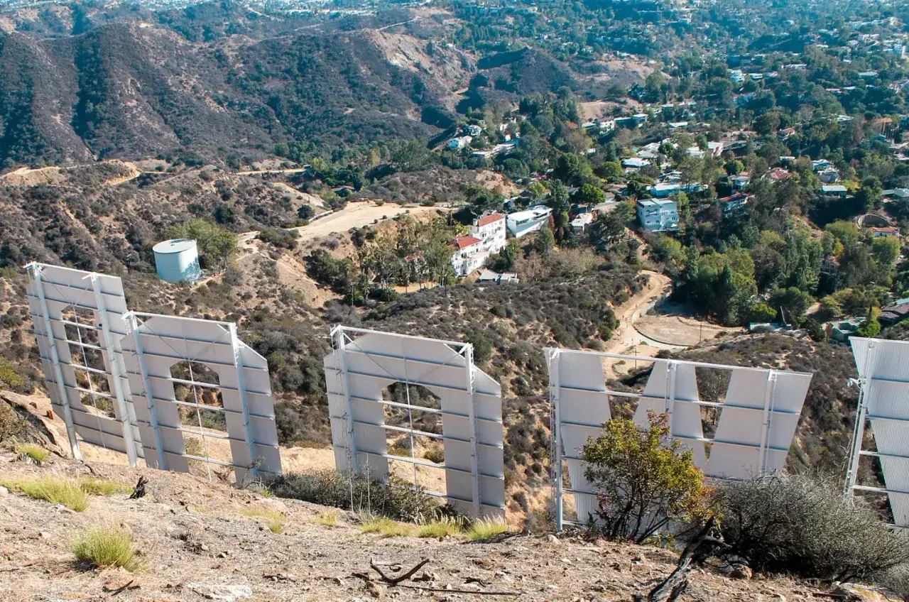 Widok zza gigantycznych liter znaku Hollywood. W oddali widać wzgórza z domami i drogami.